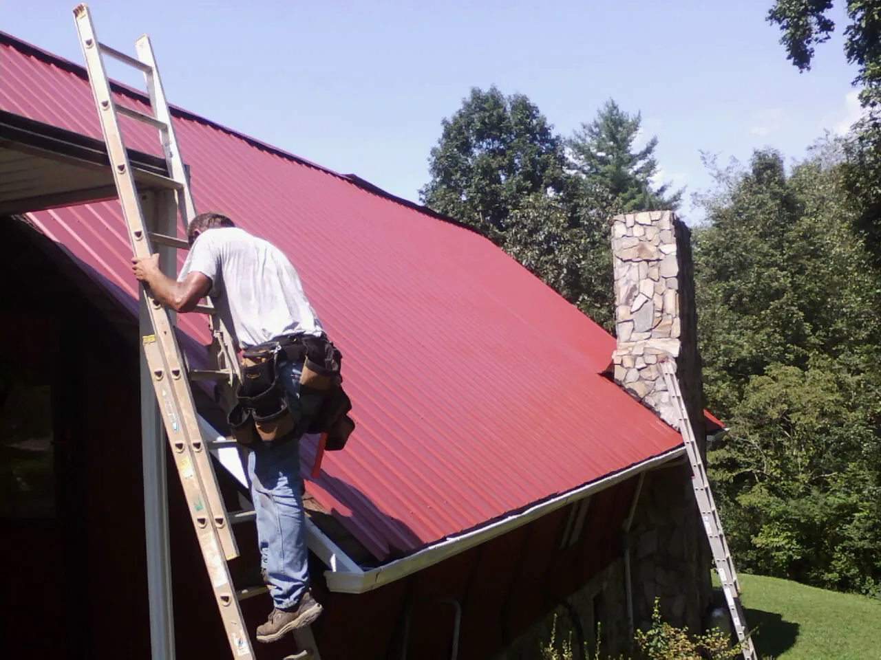 Roofer on a ladder beside a red metal roof and stone chimney — Soffit & Fascia Repair by Roofing Brothers in Minotola